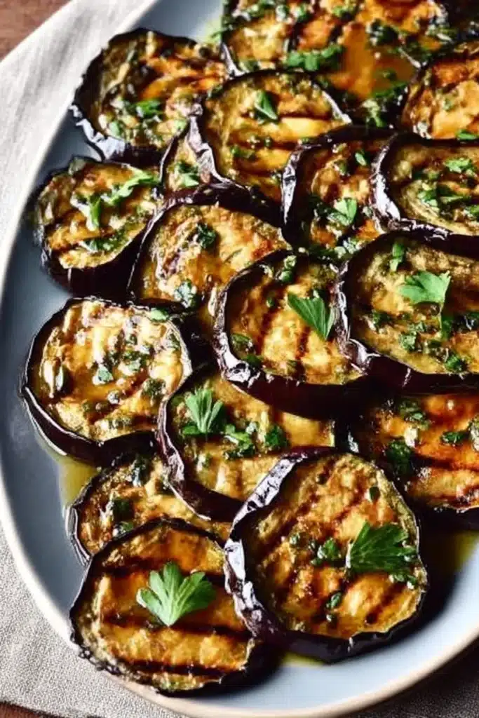 Grilled eggplant slices with garlic and herbs on a white platter, topped with fresh parsley and char marks