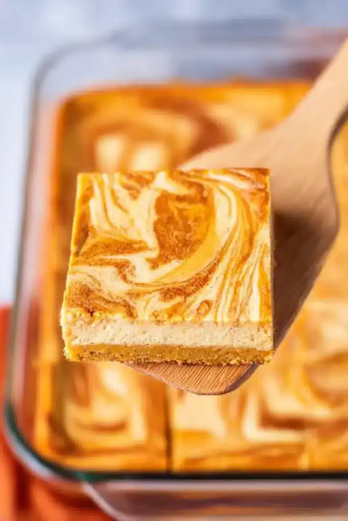 Pumpkin cheesecake bar with pumpkin swirl lifted from baking dish on wooden spatula

