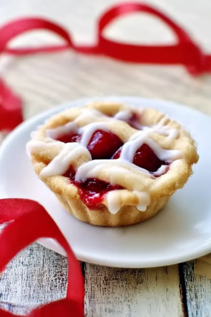 Cherry Pie Cookie Cups – Easy Bite-Sized Party Treats 5 Cherry pie cookie cup with icing and cherry filling on a white plate with red ribbon on rustic wood