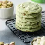Pistachio Pudding Cookies – Soft, Chewy & Easy to Make 7 Stack of soft green pistachio pudding cookies with white chocolate chips, placed on a wire rack with pistachios and baking ingredients around.