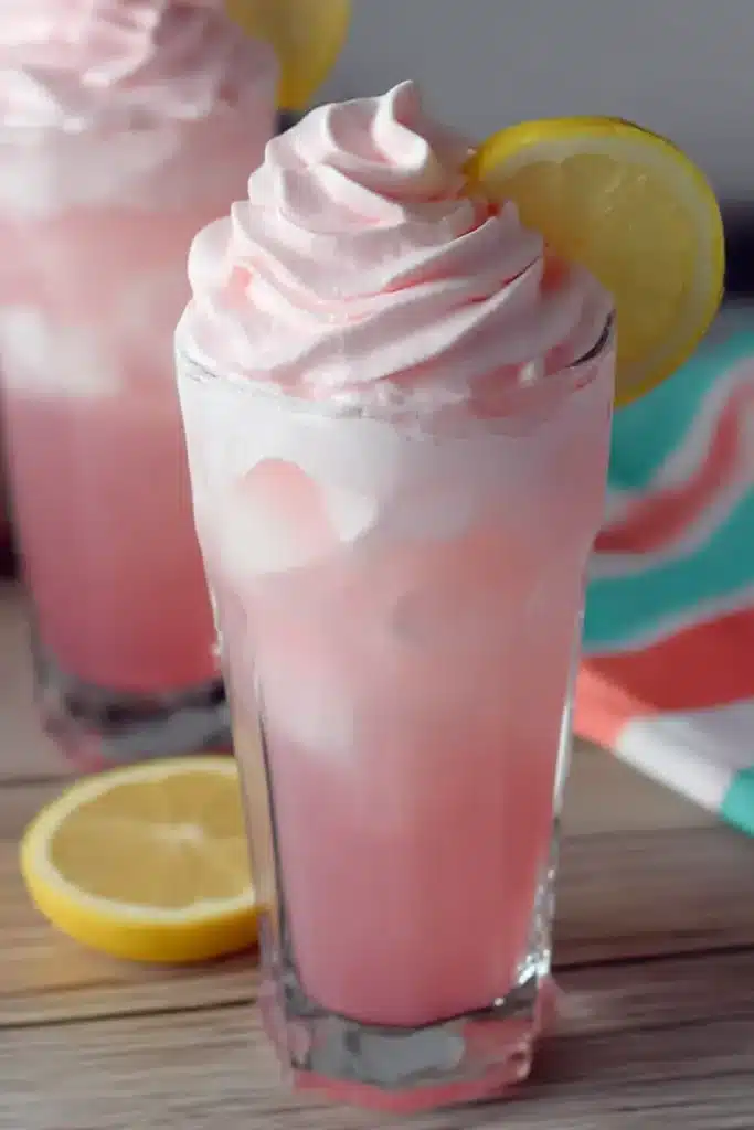 Whipped Pink Lemonade – Quick, Fun & Refreshing Favorite 8 Tall glass of whipped pink lemonade with creamy topping and lemon garnish on wood, styled with pastel striped napkins.