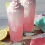 Whipped Pink Lemonade – Quick, Fun & Refreshing Favorite 9 Tall glasses of whipped pink lemonade with lemon slices and creamy pink topping on a wooden board with striped napkins.