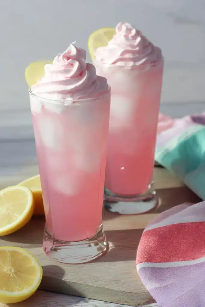 Tall glasses of whipped pink lemonade with lemon slices and creamy pink topping on a wooden board with striped napkins.