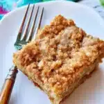 Banana Crumb Coffee Cake – Sweet, Moist & Easy to Make 12 A slice of banana crumb coffee cake with a crunchy cinnamon topping on a white plate with a fork, styled on a rustic white table.