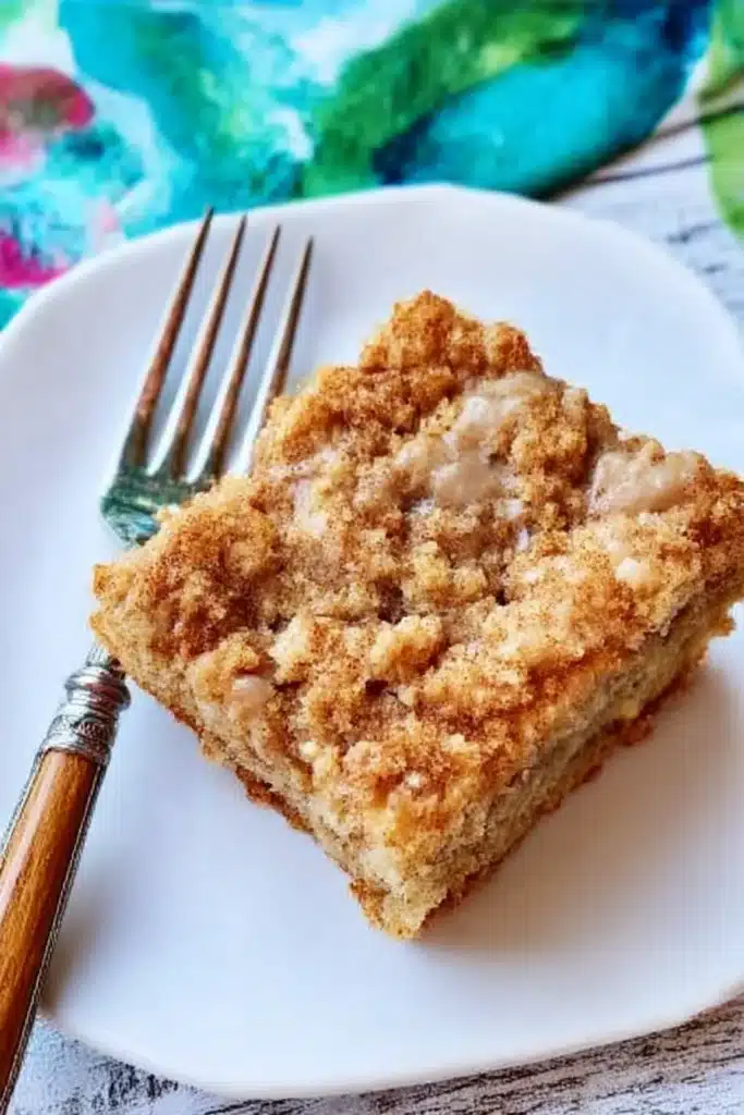 A slice of banana crumb coffee cake with a crunchy cinnamon topping on a white plate with a fork, styled on a rustic white table.