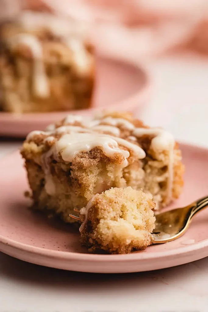 Moist banana crumb cake slice with cinnamon topping and cream cheese drizzle on a pink plate, fork holding a bite.