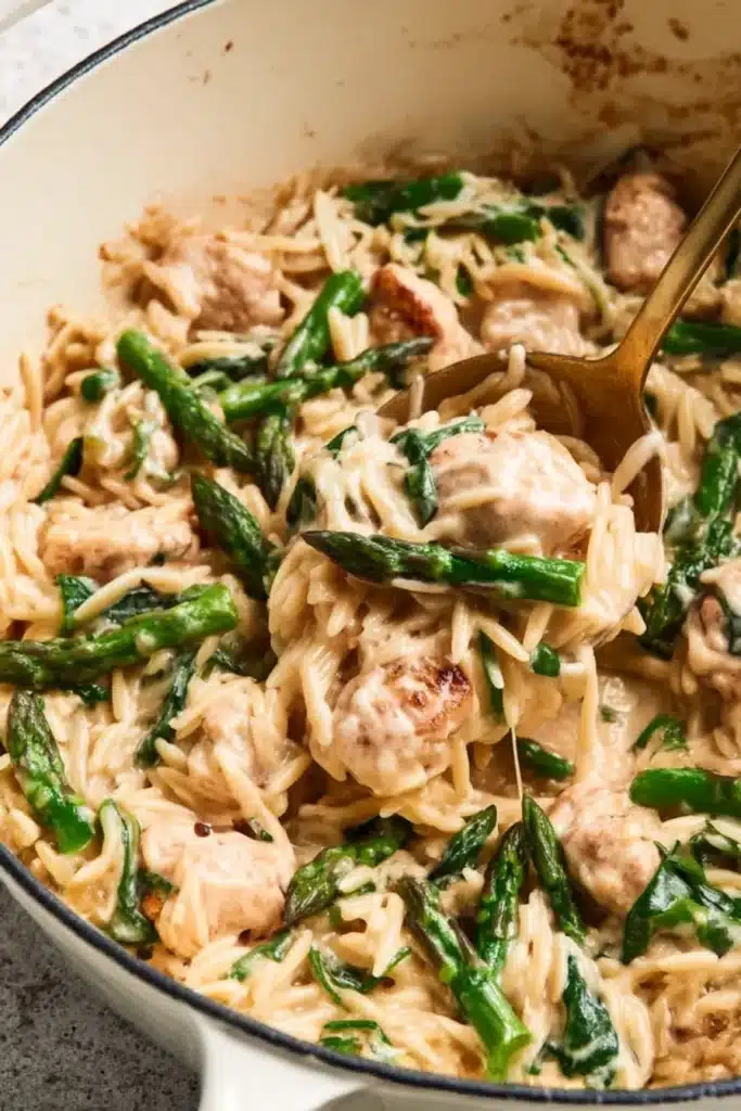 Close-up of creamy chicken orzo pasta with asparagus and parmesan in a white Dutch oven with a golden spoon