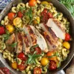 Easy Chicken Pesto Pasta 7 Sliced seared chicken breast over Easy Chicken Pesto Pasta with cherry tomatoes and fresh basil in a skillet, on a rustic tabletop with a floral plate nearby.