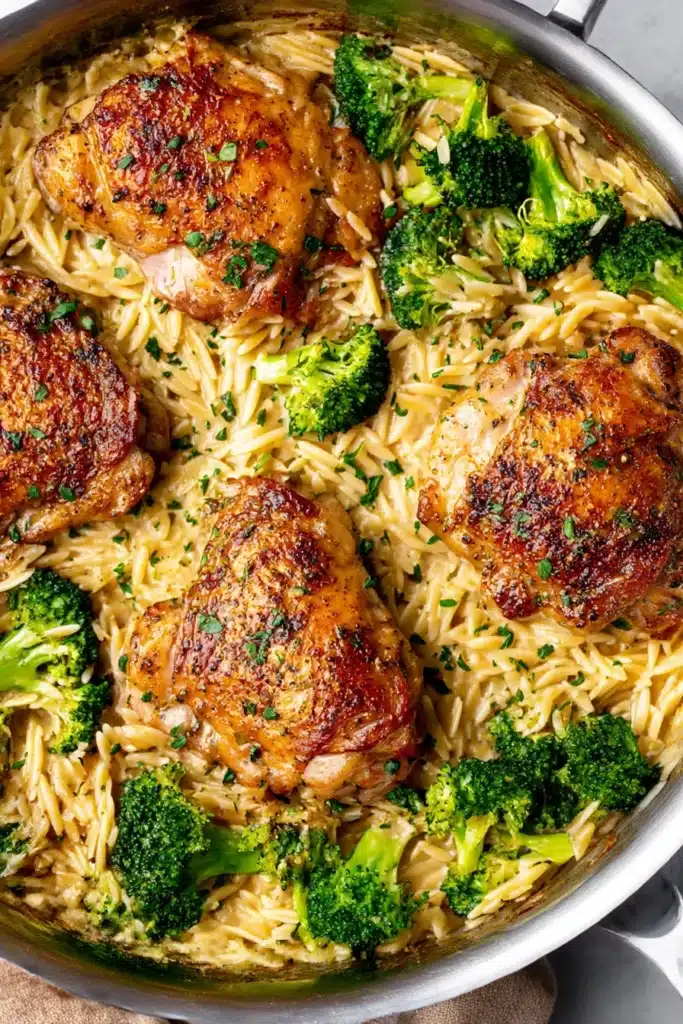 Skillet chicken thighs with creamy orzo and broccoli, topped with herbs, in a stainless steel pan on a rustic beige stone surface