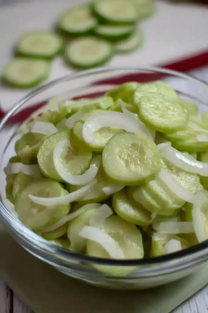 Glass bowl of cucumber and onion in vinegar with sliced cucumbers in the background