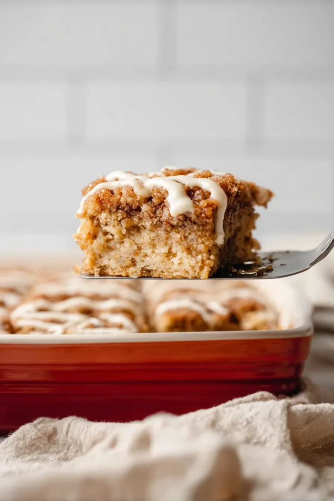 Moist banana crumb cake slice lifted on spatula with crumb topping and drizzle, red baking dish and white tile background.