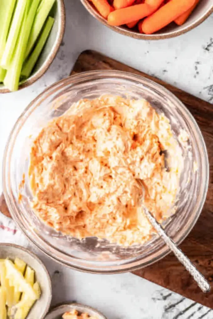 Buffalo Chicken Dip Recipe (Easy, Creamy & Crowd-Pleasing) 9 Mixed Buffalo chicken dip in glass bowl with spoon, surrounded by carrots and celery