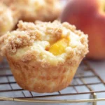 Keto Peach Cobbler Muffins 9 Keto peach cobbler muffin with streusel topping on a cooling rack, with whole peaches in background