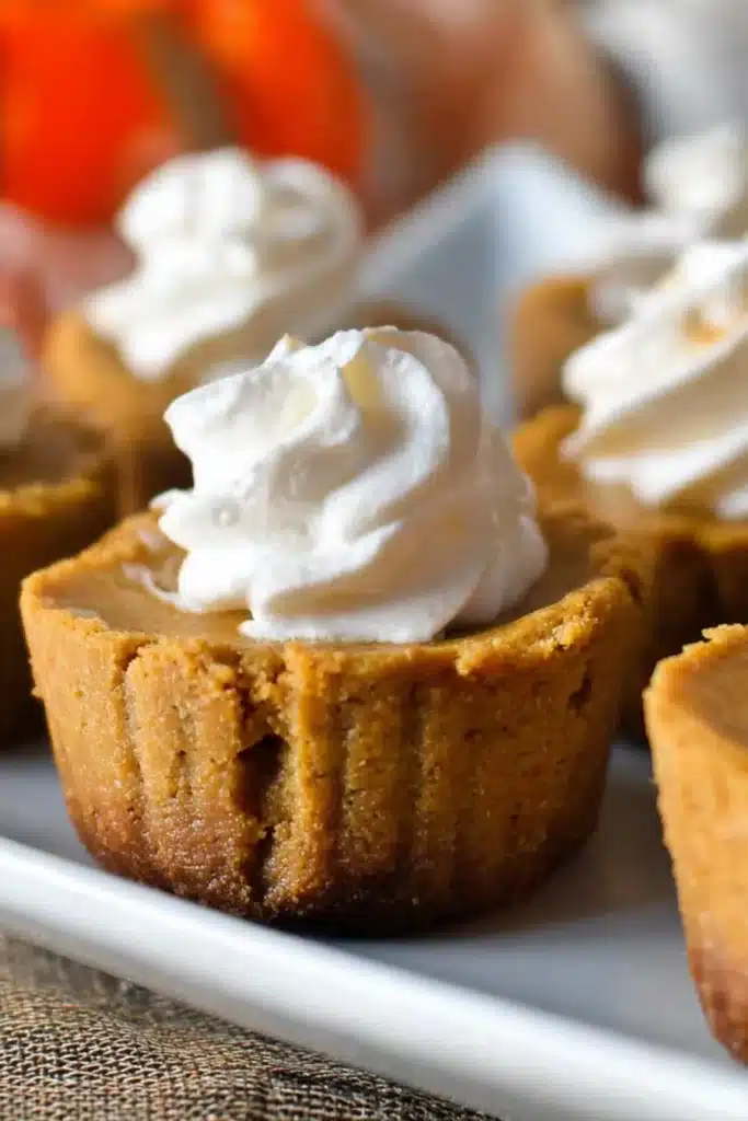 Close-up of mini keto pumpkin pies topped with whipped cream on a white platter with fall-themed background.
