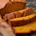 Sliced homemade pumpkin bread loaf showing golden crust and moist orange interior on rustic board.