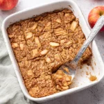 Oatmeal Apple Breakfast Bake – Cozy & Vegan Delight 7 Oatmeal apple breakfast bake in a white dish with golden crispy top, served with a silver spatula and fresh apples
