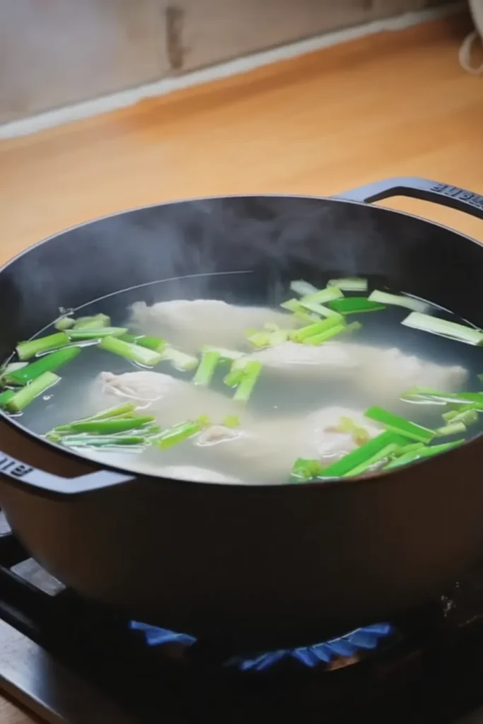 Chicken breasts poaching in a black pot with green scallions in simmering broth on a light wooden table