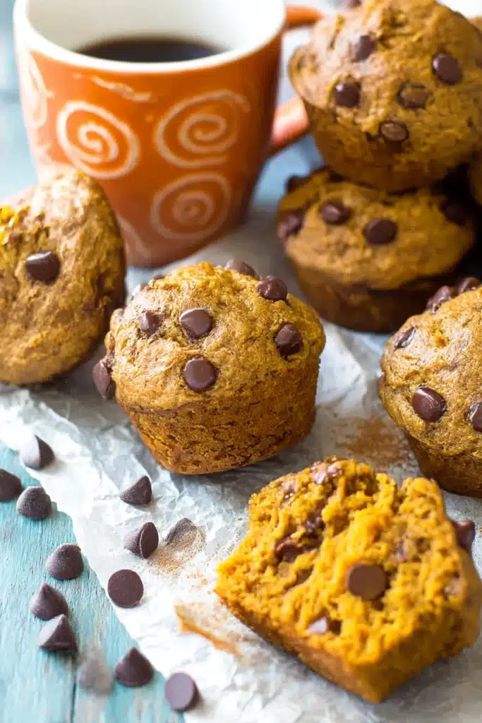 Pumpkin Chocolate Chip Muffins 9 Rustic pumpkin chocolate chip muffins on parchment with broken muffin showing moist interior, chocolate chips, and cinnamon dusting.