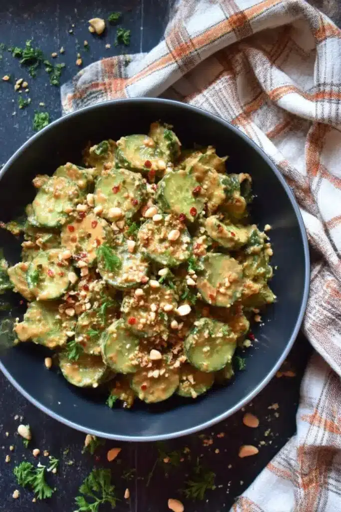 Wide overhead view of cucumber salad in spicy peanut sauce with chopped peanuts and herbs on dark tile with plaid napkin
