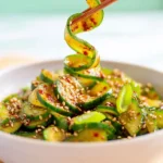 Korean Cucumber Salad – Spicy, Crunchy Summer Side 7 Spiralized Korean cucumber salad with chili dressing in a white bowl lifted by chopsticks