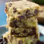 Chocolate Chip Zucchini Bars – Simple & Delicious Treat 5 Four chocolate chip zucchini bars stacked on a white plate with fresh zucchini and chocolate chips in the background