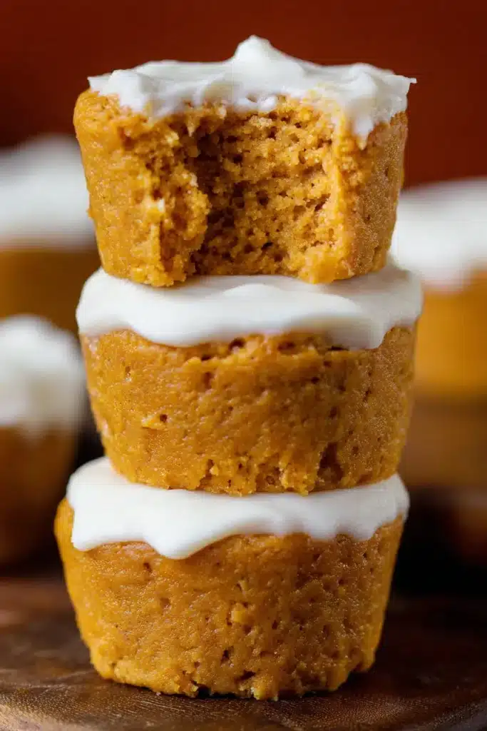 Stack of flourless pumpkin muffins with white frosting, top muffin with bite showing moist crumb