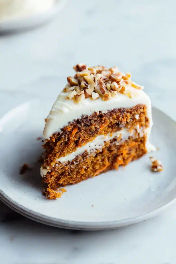 Slice of easy carrot cake with frosting and chopped pecans on a white plate