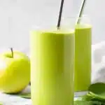 Refreshing spinach green apple smoothie in tall glasses with metal straws, fresh apple and spinach leaves on wooden table