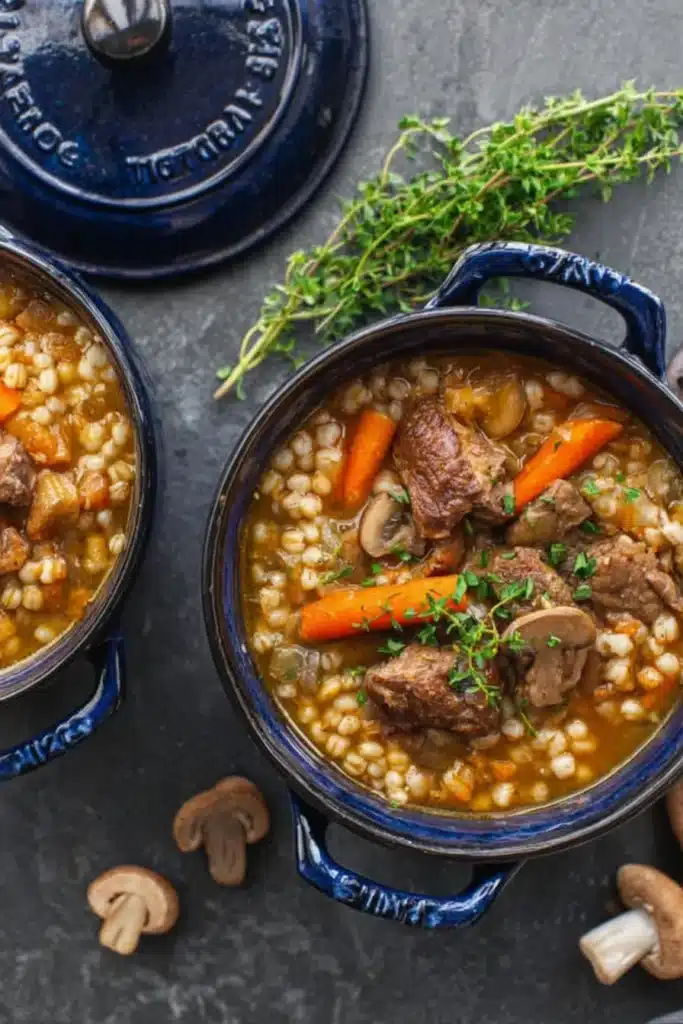 Two bowls of beef barley soup with carrots, mushrooms, and herbs