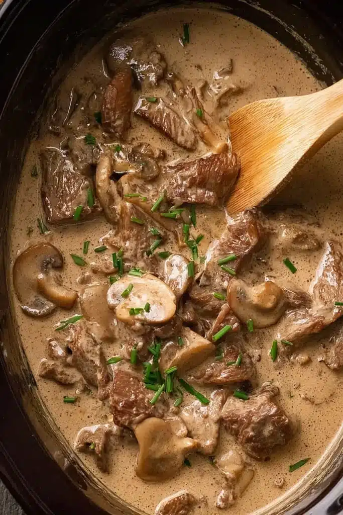 Beef stroganoff stew cooking in a pot with wooden spatula and fresh herbs