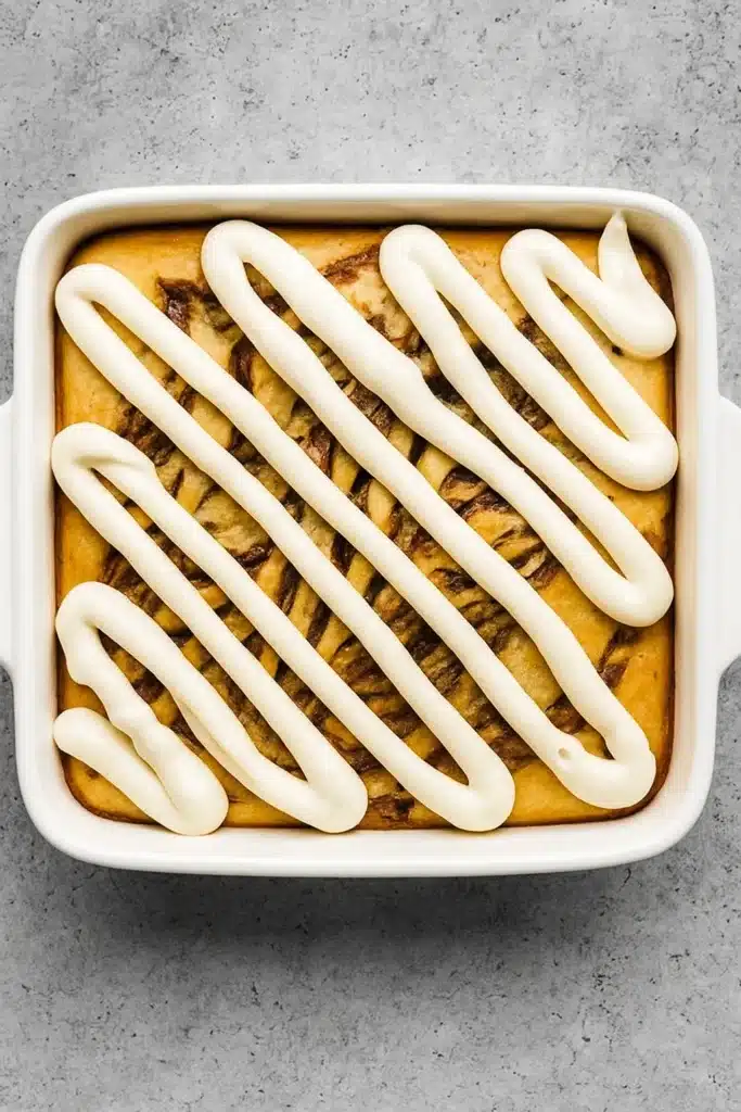 Baked cinnamon roll cake with cream cheese frosting in white square dish