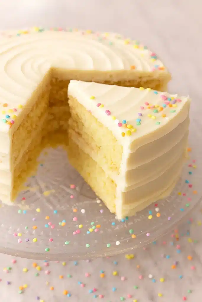 Vanilla cake recipe with buttercream frosting and colorful sprinkles slice on glass stand