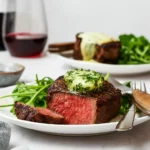 A perfectly cooked filet mignon with compound butter and fresh arugula on a white plate, paired with red wine.