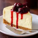 Slice of perfect cheesecake with cherry topping and graham crust on white plate