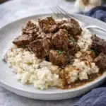 Crockpot steak bites with gravy over mashed potatoes on a white plate