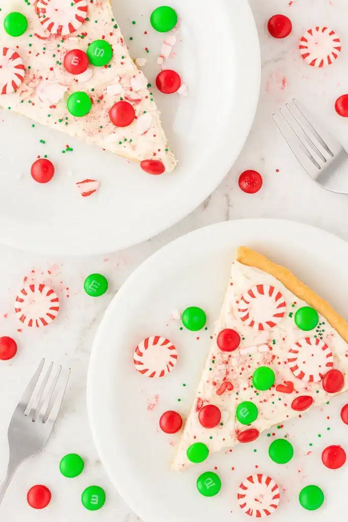Two slices of Christmas cookie pizza with frosting, candies, and peppermint on white plates