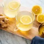 Two glass cups of ginger lemon tea with fresh ginger and lemon on a marble table