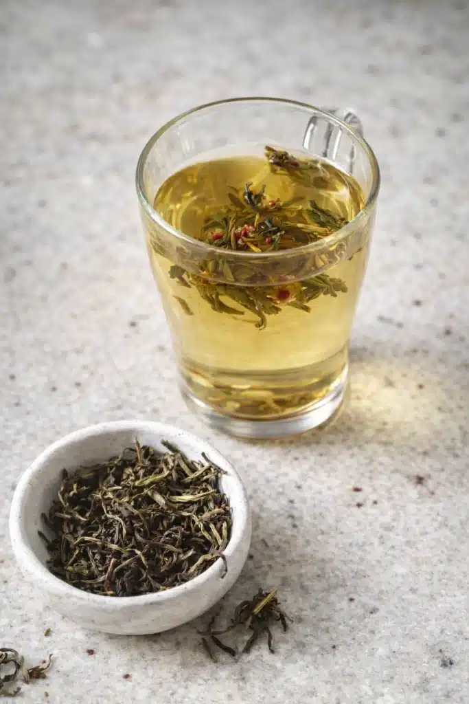 Glass of green tea with floating leaves beside bowl of loose tea on white table