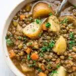 Warm bowl of lentil potato soup with carrots and parsley on light surface
