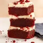 Fudgy red velvet brownies stacked with cream cheese frosting and red crumbs