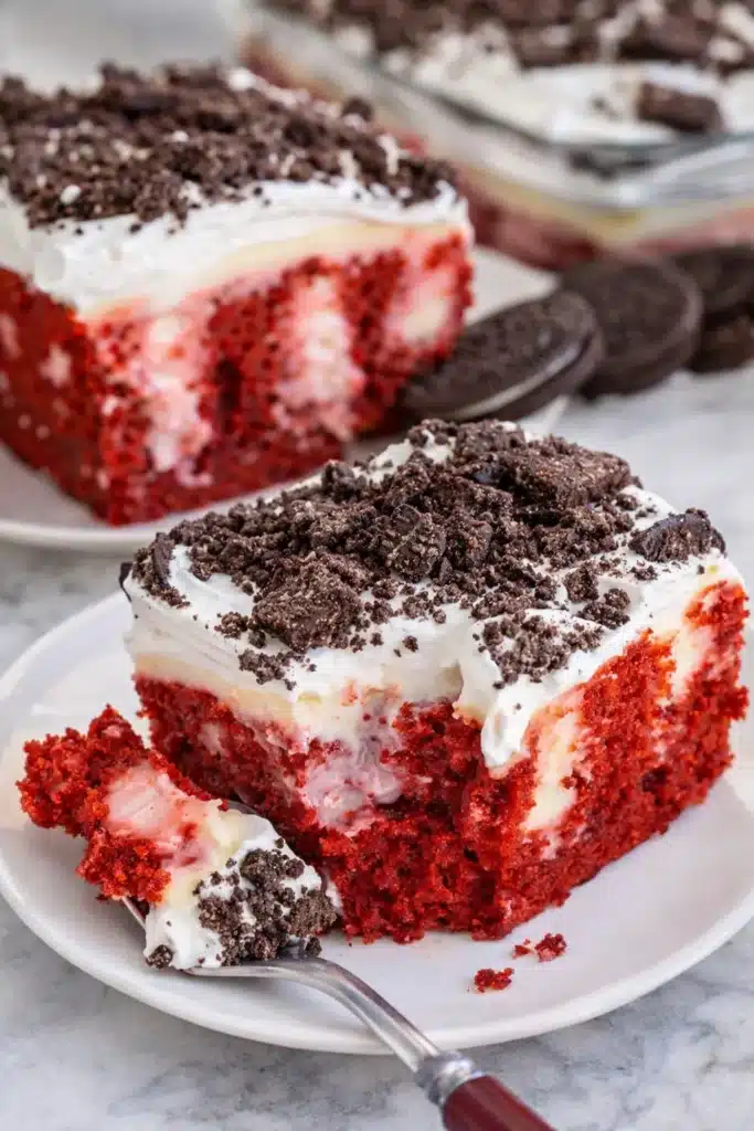 Slice of red velvet poke cake with whipped topping and crushed Oreos on marble surface