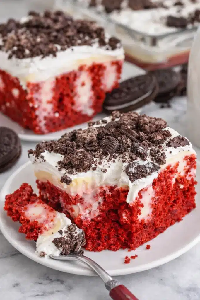 Red velvet poke cake slice with whipped topping and crushed Oreos on white plate