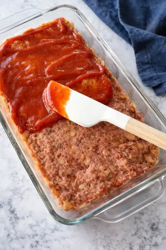 Raw meatloaf in glass pan topped with ketchup glaze and spatula