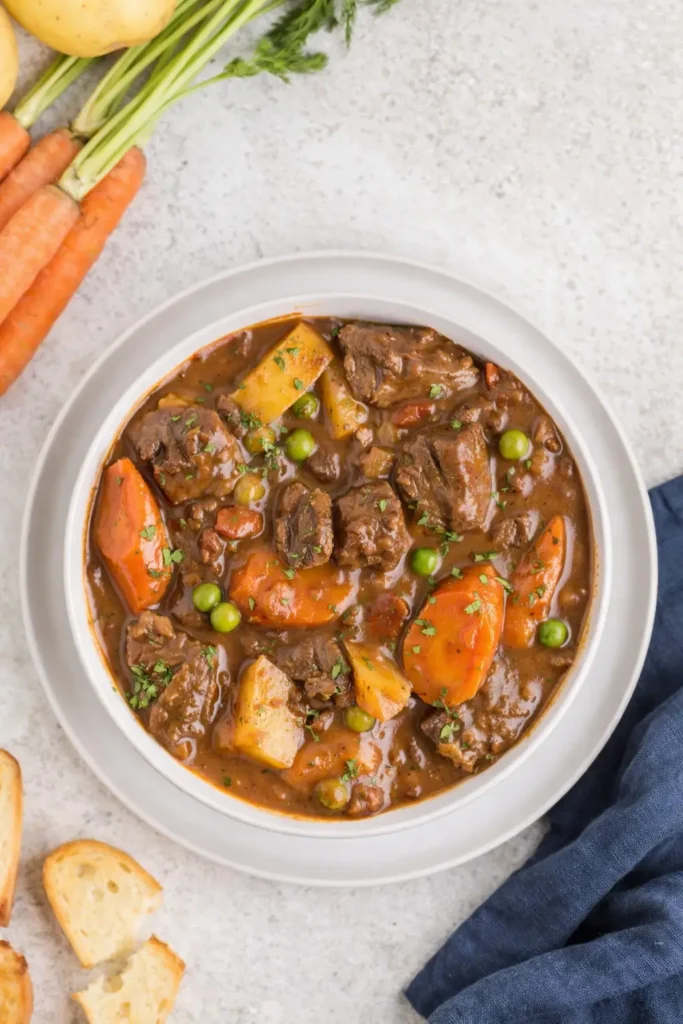 Bowl of Dutch oven beef stew with carrots, potatoes, and peas beside a dark blue napkin