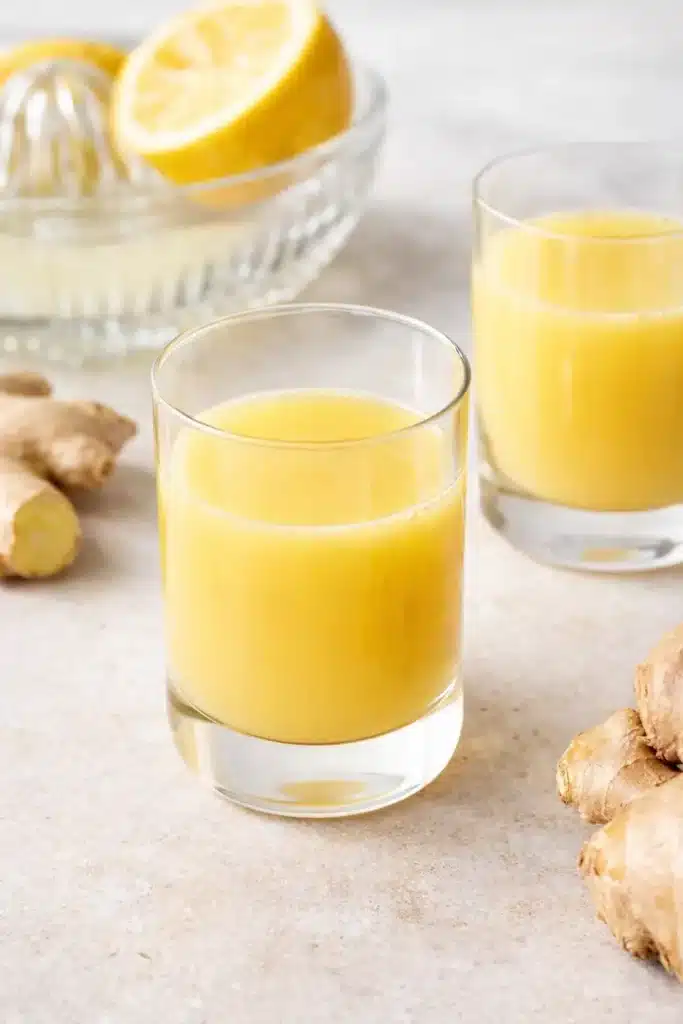 Two glasses of fresh ginger shots with lemon and ginger root on a light stone surface