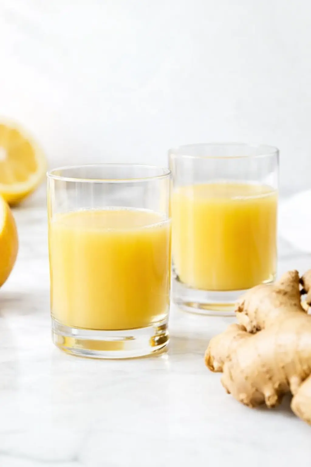 Two glasses of fresh ginger shots with lemon and ginger root on a bright countertop