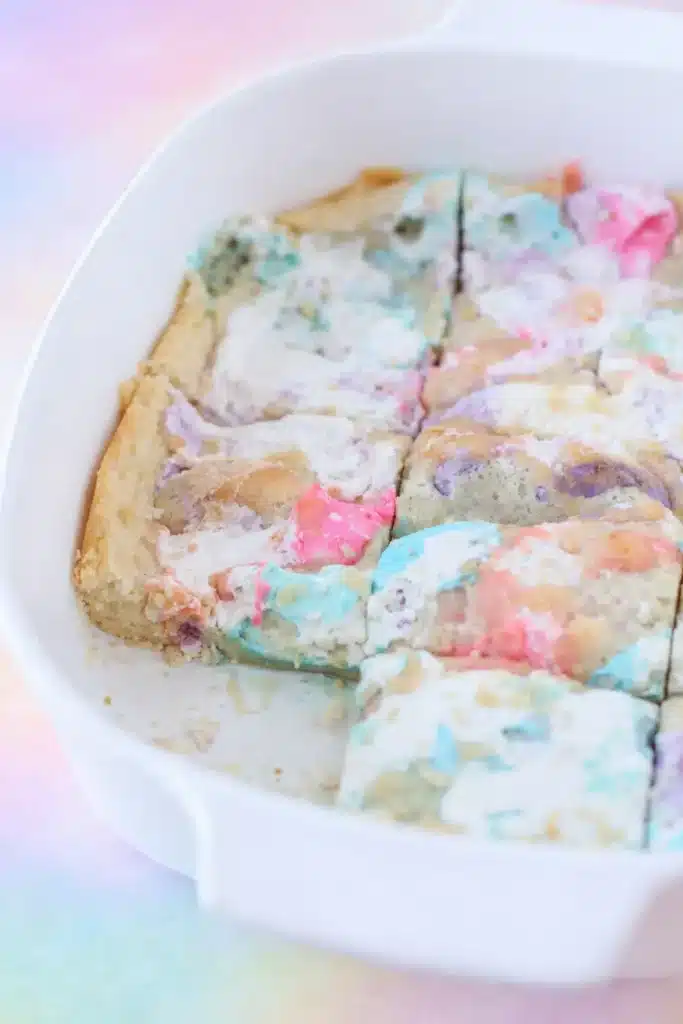 Easter Peeps Sugar Cookie Bars Recipe 12 Peeps sugar cookie bars partially sliced in a white dish with pastel melted marshmallow swirls.