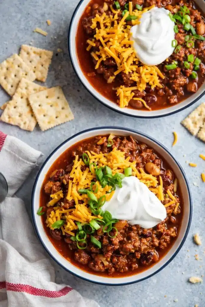 Two bowls of homemade chili topped with cheese, sour cream, and green onions