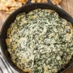 Creamy spinach artichoke dip in skillet with pita chips on wooden board