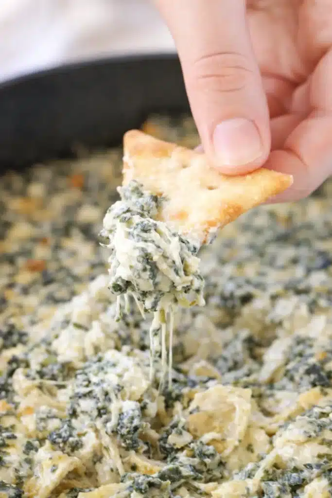 Hand dipping cracker into cheesy spinach artichoke dip
